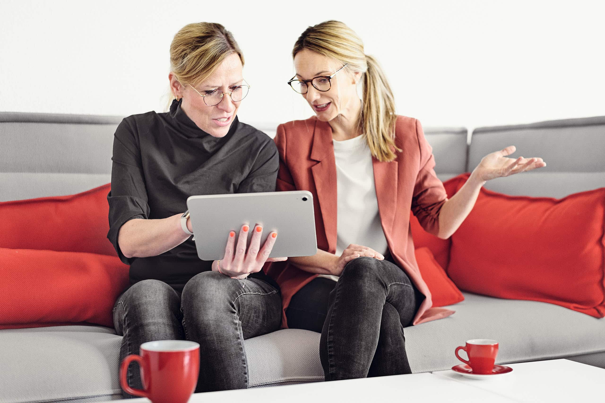 Magdalena Sabaß explique quelque chose à Céline Spitz sur une tablette, alors qu'elles sont assises sur un canapé et qu'un café est posé sur la table basse.