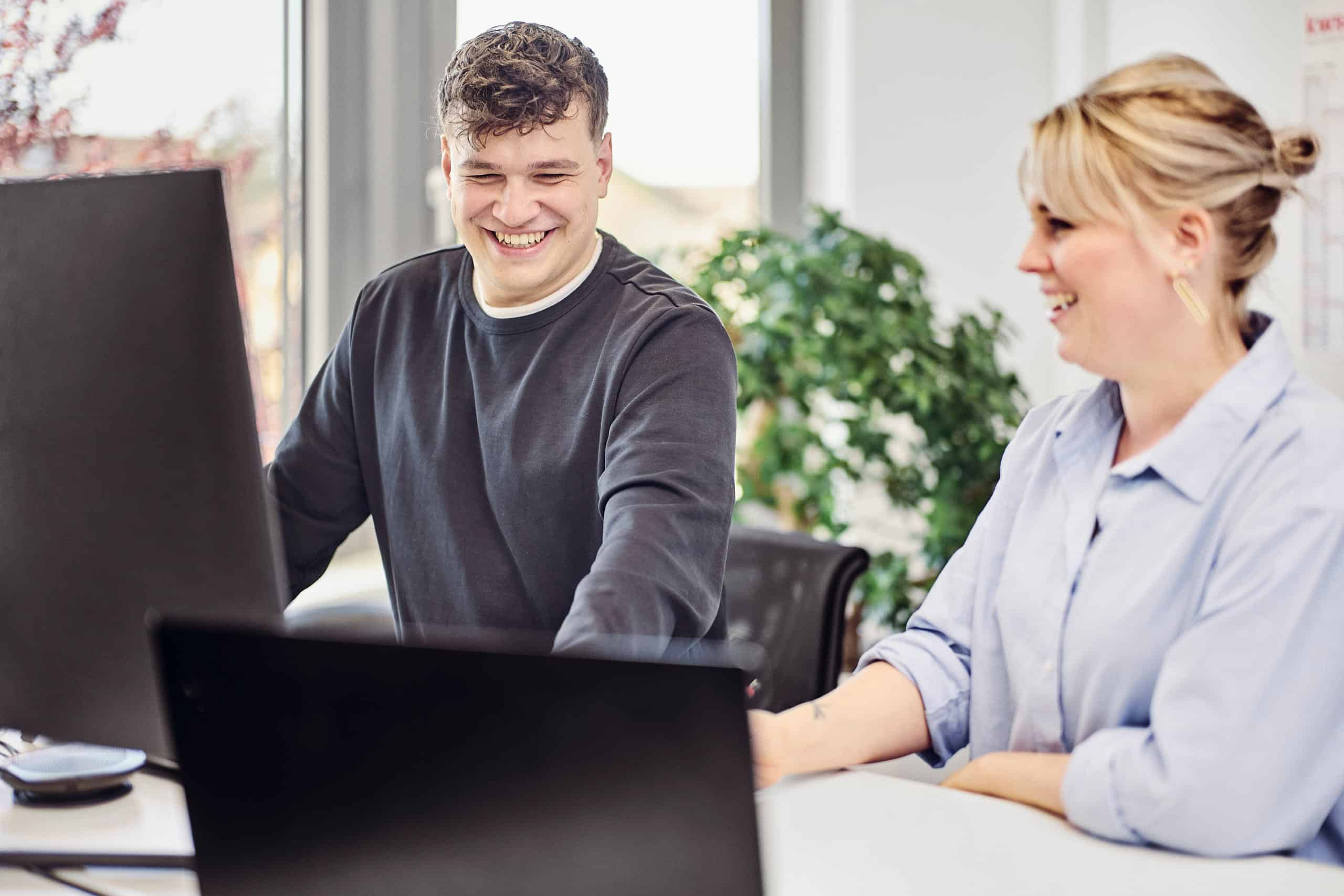 Julius Binninger et Anna Wißler se tiennent debout devant le bureau, le sourire aux lèvres, les yeux rivés sur leurs écrans.