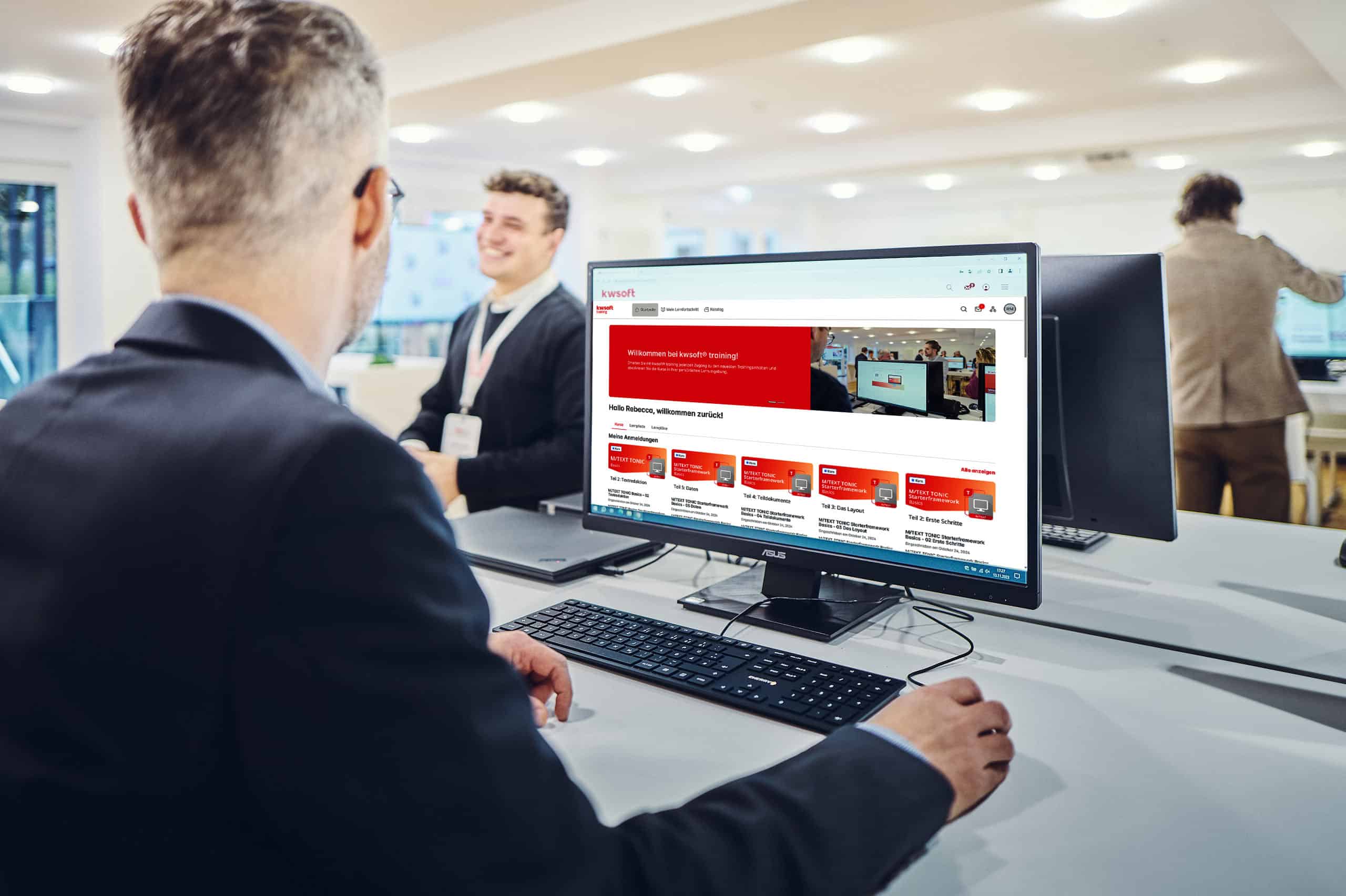 Un homme est assis devant un écran sur lequel est affichée une formation kwsoft® et regarde un homme debout à l'arrière-plan.